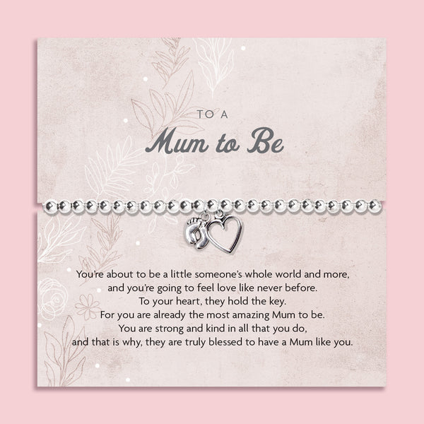 A silver beaded bracelet with a small heart and footprint charm, presented on a personalized 'Mum to Be' card with a quote, set against a solid pink background.