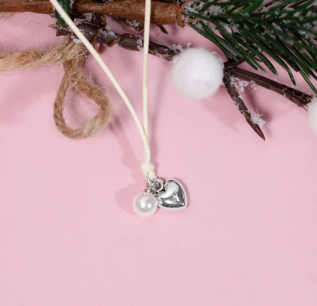 A delicate bracelet with a white cord, a small white pearl bead, and a silver heart charm, displayed against a soft pink background with rustic holiday props.
