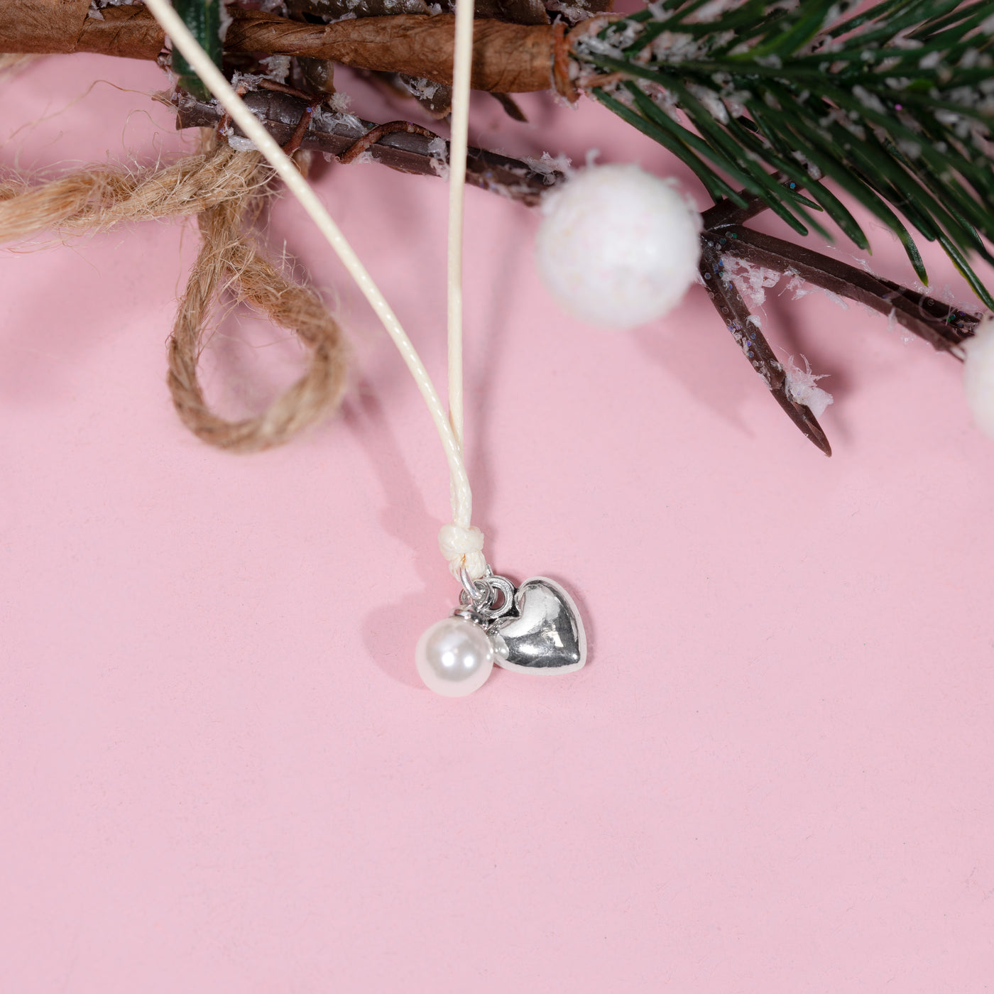 A delicate bracelet with a white cord, a small white pearl bead, and a silver heart charm, displayed against a soft pink background with rustic holiday props.