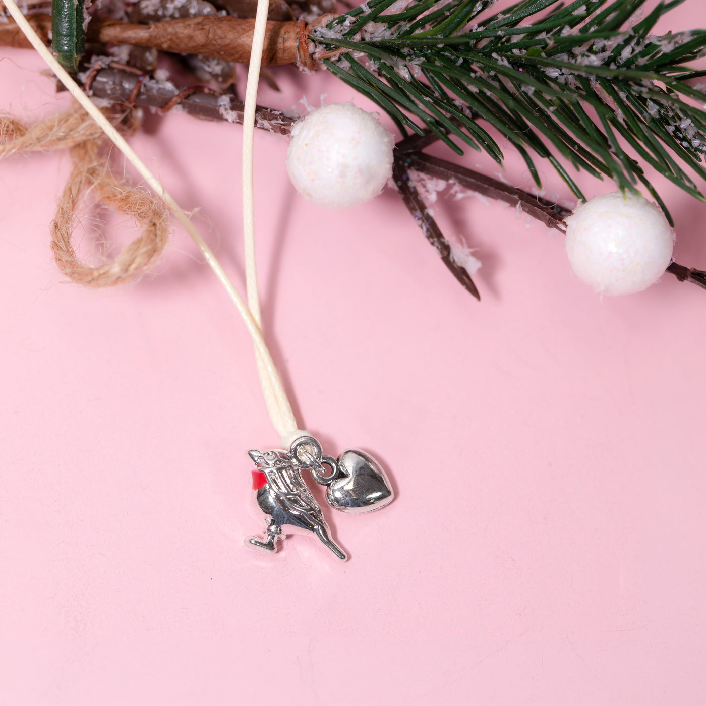 A silver metal Christmas bracelet with robin and heart charms, hanging from a cream cord against a pink background with decorative pine branches, white pom-poms, and twine.