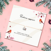 A Christmas-themed card with a thin adjustable bracelet, featuring robin illustrations and the text 'Robins appear' and a poem. The card is styled on a pink background with pine branches and ornaments.