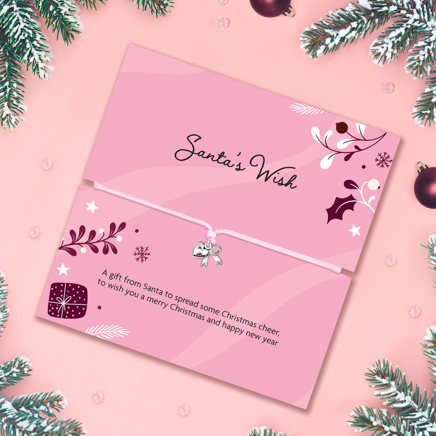 A pink Christmas card with a small silver bell charm on a white string, shown open on a pink background with pine branches and ornaments around the edges. The card reads 'Santa's Wish' and includes a holiday message.