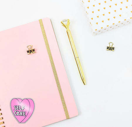 Pink notebook with gold accents, a gold pen, and a 'Self Care' heart sticker on a white background.