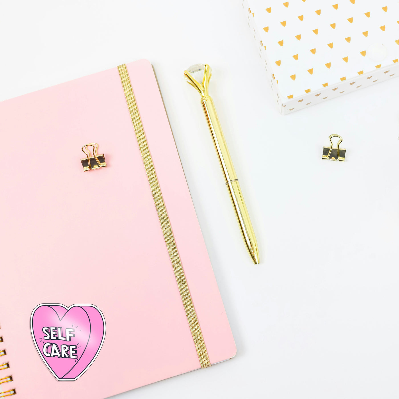 Pink notebook with gold accents, a gold pen, and a 'Self Care' heart sticker on a white background.
