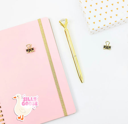 Pink notebook with gold accents and a gold pen on a white background, featuring 'Silly Goose' sticker.