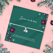 A Christmas-themed gift card featuring a delicate white cord bracelet with a silver heart charm and pearl bead. The card is dark green with festive illustrations of snowflakes, wrapped presents, and pine branches, and is styled on a pink background with holiday decorations.