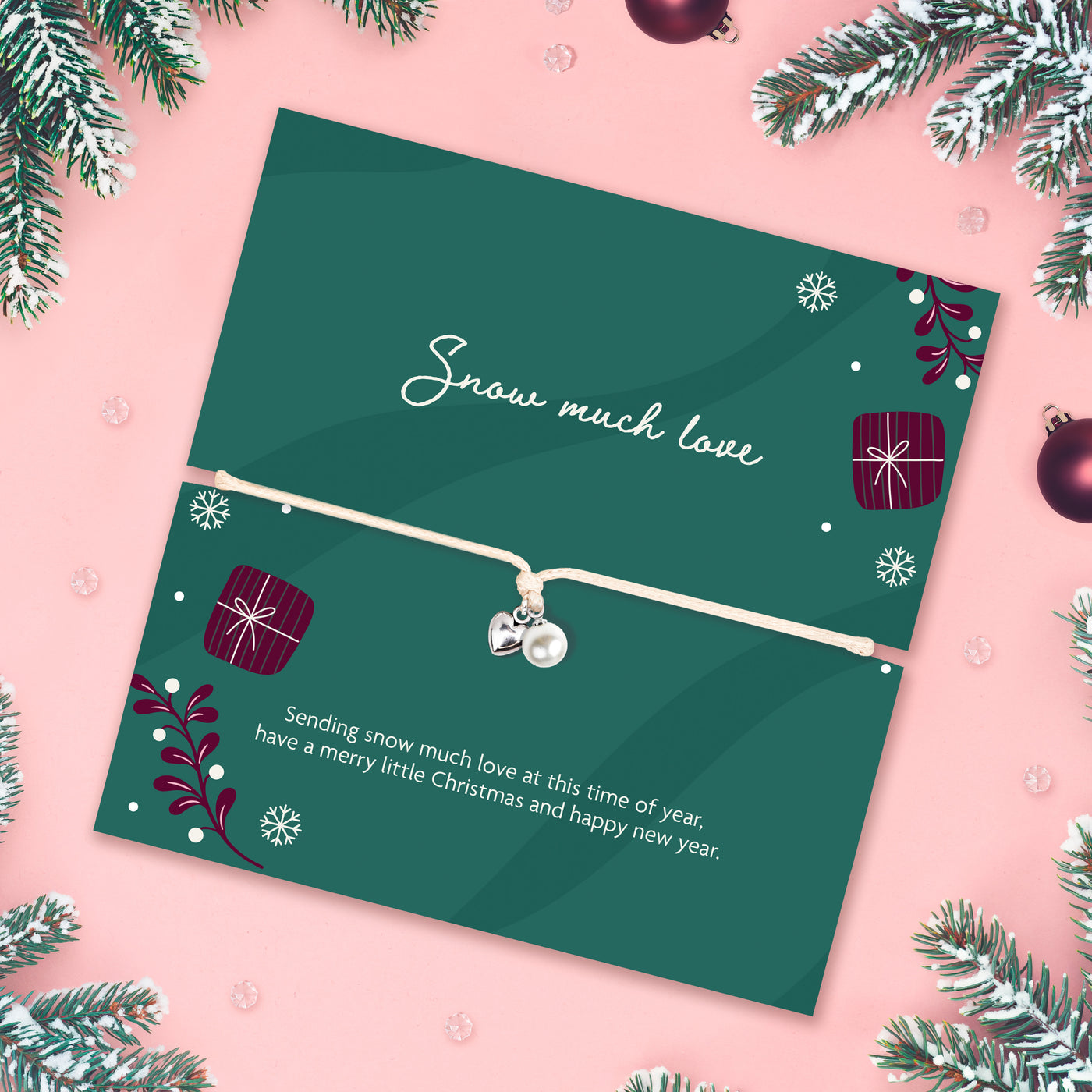A Christmas-themed gift card featuring a delicate white cord bracelet with a silver heart charm and pearl bead. The card is dark green with festive illustrations of snowflakes, wrapped presents, and pine branches, and is styled on a pink background with holiday decorations.