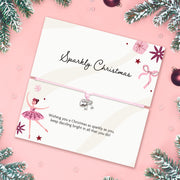 A pink cord bracelet with a small silver bow charm is displayed on a white card that reads 'Sparkly Christmas' with holiday illustrations, set on a pink background with pine branches and ornaments.