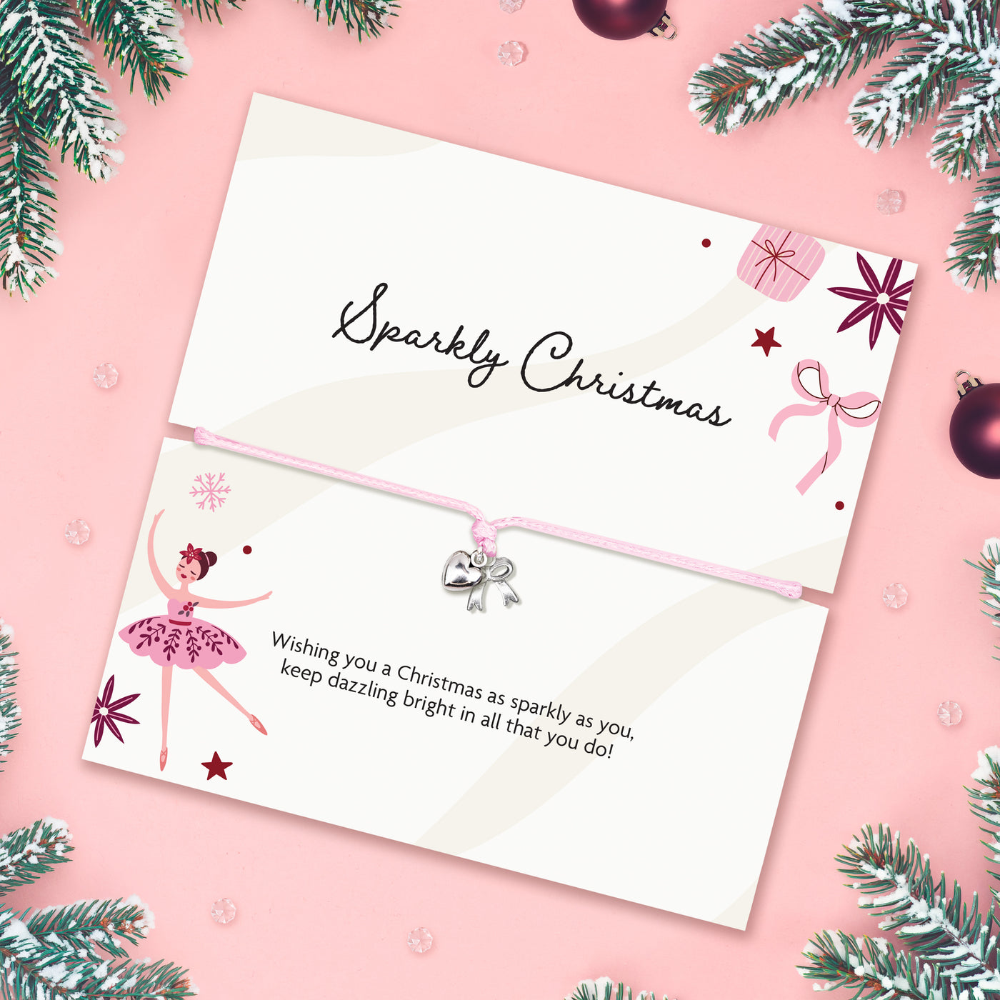 A pink cord bracelet with a small silver bow charm is displayed on a white card that reads 'Sparkly Christmas' with holiday illustrations, set on a pink background with pine branches and ornaments.