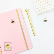 Pink notebook with gold accents, a gold pen, and decorative clips on a white background. With a retro stamp sticker on the journal.