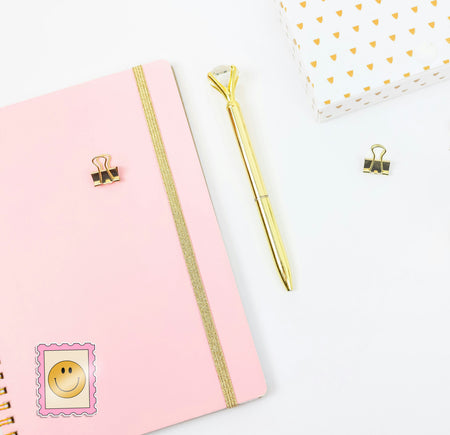 Pink notebook with gold accents, a gold pen, and decorative clips on a white background. With a retro stamp sticker on the journal.
