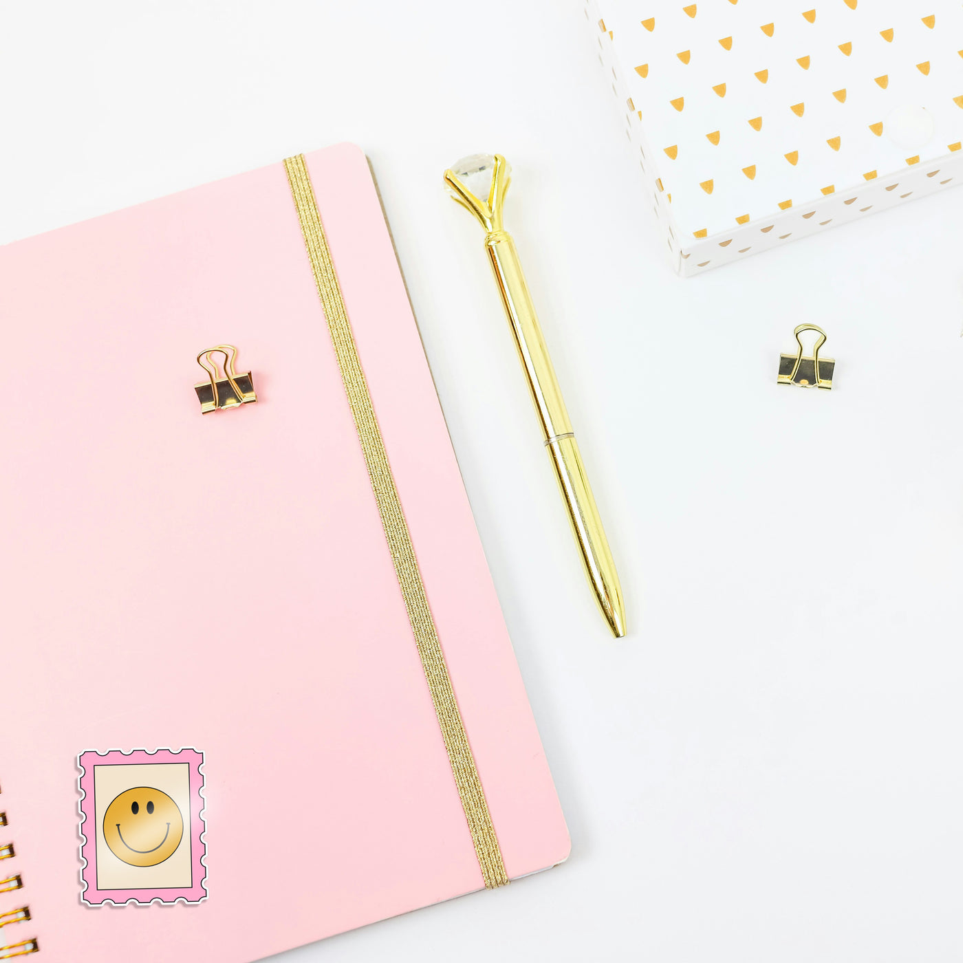 Pink notebook with gold accents, a gold pen, and decorative clips on a white background. With a retro stamp sticker on the journal.