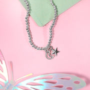 A silver beaded bracelet with a crescent moon and star charms, styled on a pink background with a light green card and a butterfly cutout.