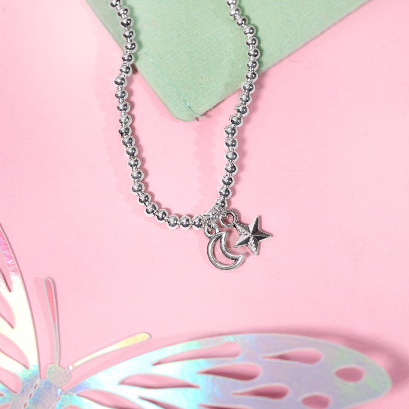 A silver beaded bracelet with a crescent moon and star charms, styled on a pink background with a light green card and a butterfly cutout.