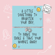 A silver charm bracelet displayed on a light blue cards with handwritten-style positive quote, set against a pink background with simple doodle illustrations.