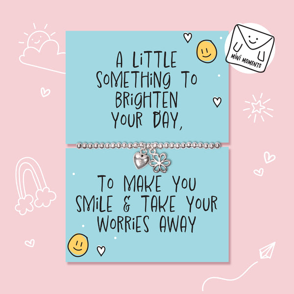 A silver charm bracelet displayed on a light blue cards with handwritten-style positive quote, set against a pink background with simple doodle illustrations.