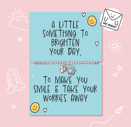A silver charm bracelet displayed on a light blue cards with handwritten-style positive quote, set against a pink background with simple doodle illustrations.