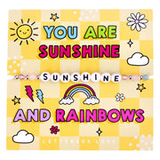 An image of a motivational card featuring a beaded bracelet with white letter beads spelling "SUNSHINE." The card has a bright yellow checkered background with bold, colorful text that reads: "You are sunshine and rainbows." The design includes playful illustrations of a smiling sun, a rainbow, clouds, flowers, and lightning bolts, creating a cheerful and vibrant theme. A fun and positive gift designed to spread happiness and remind the recipient that they bring sunshine and joy to the world.