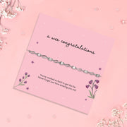 A delicate silver chain bracelet with clear rhinestones, presented on a pink card that reads 'a wee congratulations' with floral illustrations, set against a pink background with scattered baby's breath flowers.