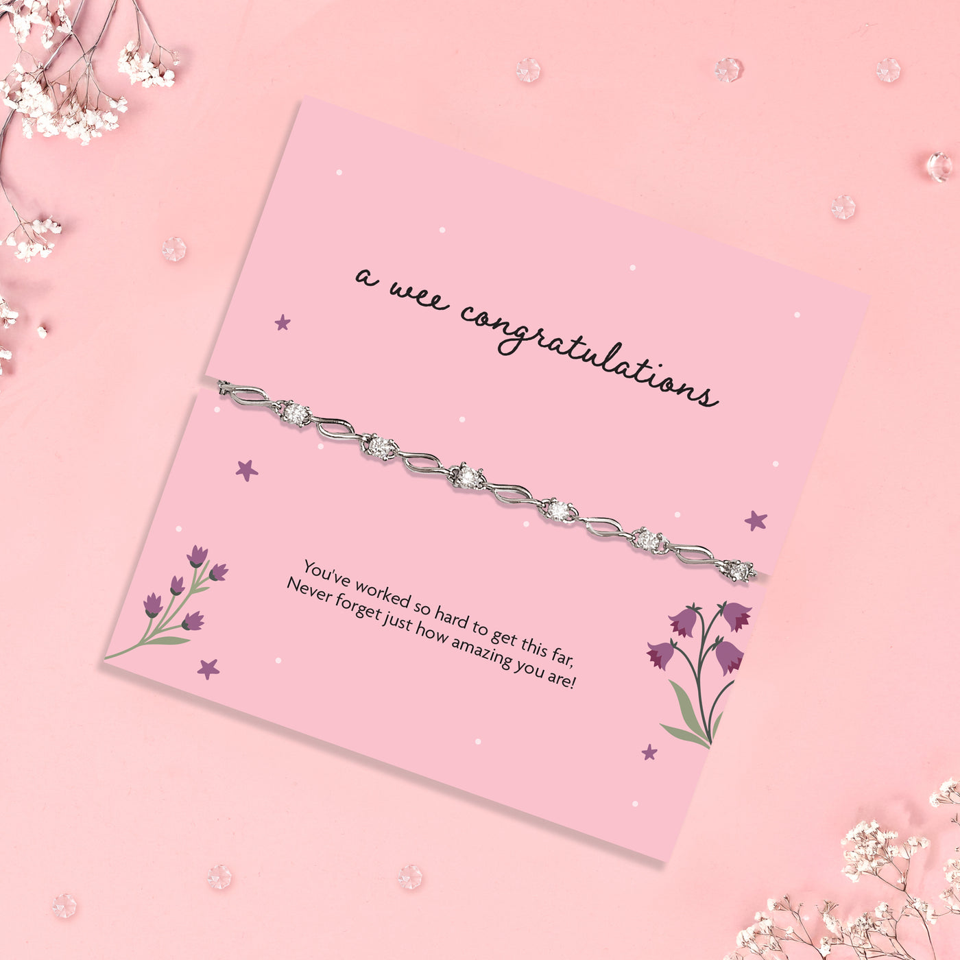 A delicate silver chain bracelet with clear rhinestones, presented on a pink card that reads 'a wee congratulations' with floral illustrations, set against a pink background with scattered baby's breath flowers.