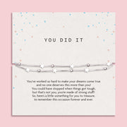 Motivational card titled "YOU DID IT" featuring a delicate silver-plated double chain bracelet with star and bead charms. The background is a soft cream tone with pastel heart confetti scattered around. A heartfelt message below the bracelet celebrates perseverance and achievement, making it a meaningful gift to mark a special milestone or success.