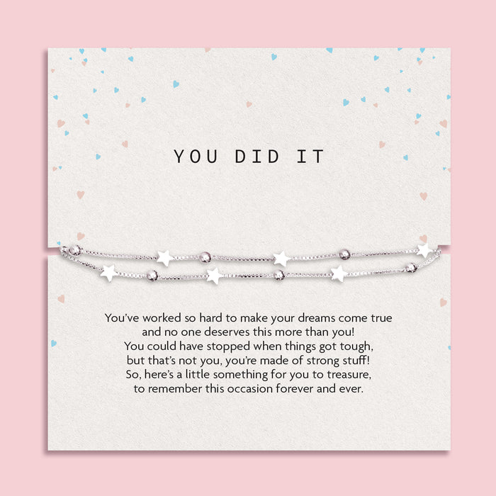 Motivational card titled "YOU DID IT" featuring a delicate silver-plated double chain bracelet with star and bead charms. The background is a soft cream tone with pastel heart confetti scattered around. A heartfelt message below the bracelet celebrates perseverance and achievement, making it a meaningful gift to mark a special milestone or success.