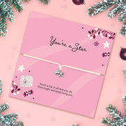 A pink card with a message of love and festive wishes and a small silver star  and heart charm on a string, styled on a pink background with pine branches and ornaments.