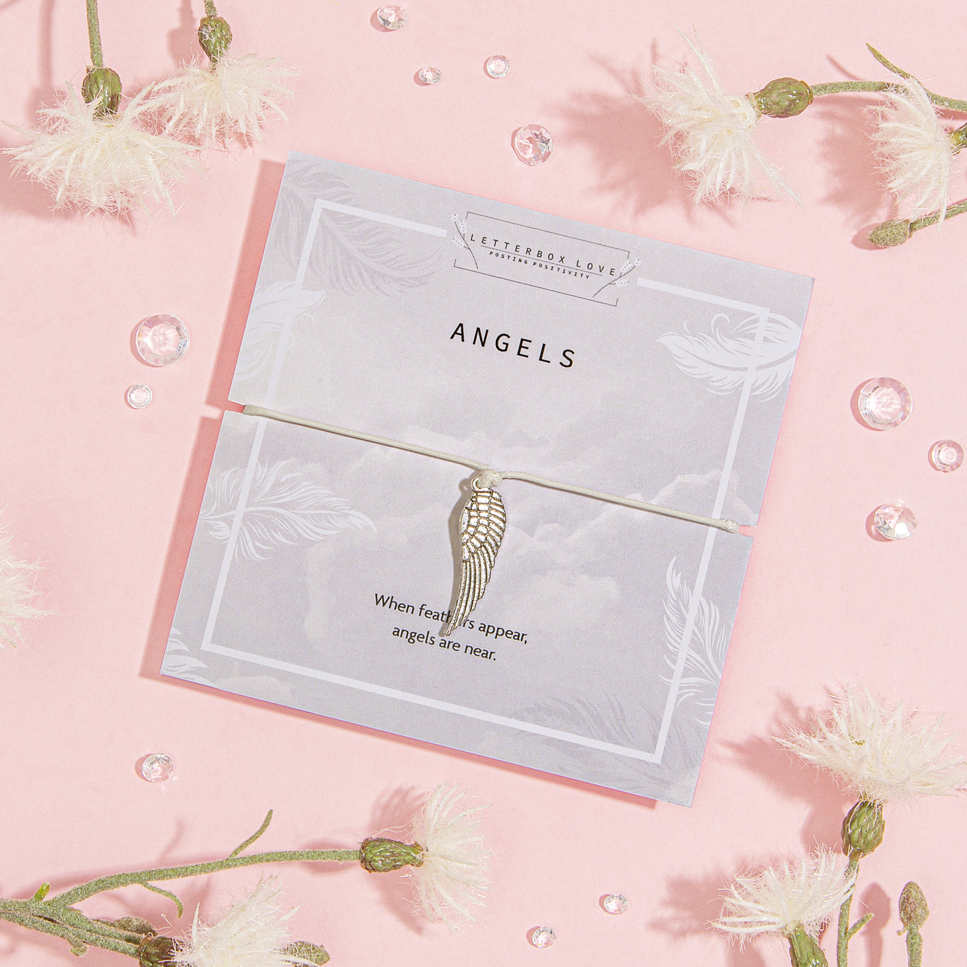 Elegant 'Angels' card on a soft pink background, decorated with subtle feather illustrations and the comforting quote, 'When feathers appear, angels are near.' Paired with the card is a delicate bracelet featuring a detailed angel wing charm. 