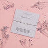 COUSIN LOVE' bracelet with a star-shaped charm, displayed on a grey card with a touching message celebrating the bond between cousins, accompanied by purple dried blossoms and scattered crystal gems on a soft pink background