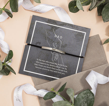 Black braided bracelet with a silver 'Dad' engraved plate, displayed on a textured gray card adorned with subtle hand silhouettes and the word 'DAD'. The card carries a touching message expressing love and admiration for a father