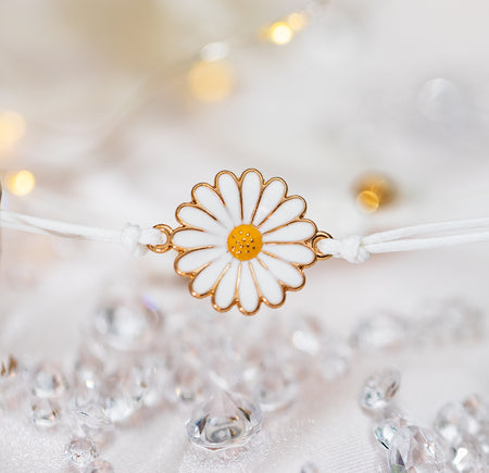 A white background with scattered gems and a cute daisy charm with gold edging on a white adjustable bracelet. 