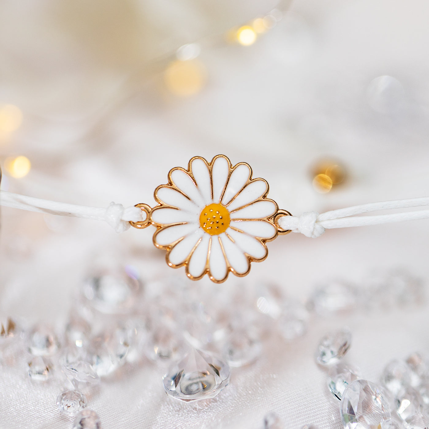 A white background with scattered gems and a cute daisy charm with gold edging on a white adjustable bracelet. 