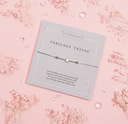 FABULOUS FRIEND' bracelet with a star-shaped charm, presented on a grey card with a heartfelt friendship message, surrounded by delicate pink blossoms and sparkling gems on a pastel pink background