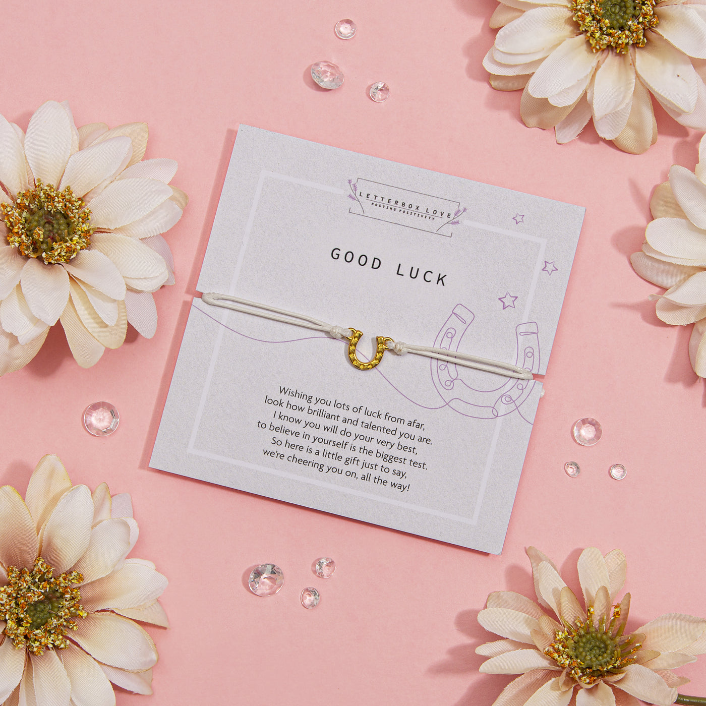 Charming 'GOOD LUCK' card on a soft pink background, adorned with encouraging words and a horseshoe illustration, accompanied by a slender silver bracelet with a golden horseshoe charm.