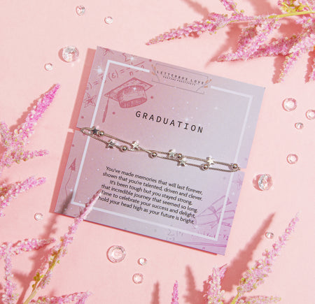 GRADUATION' bracelet with star charms and a solitary pearl, displayed on a soft pink card adorned with illustrations of a graduation cap, arrows, and math symbols. The card carries an uplifting message celebrating a graduate's achievements and bright future.