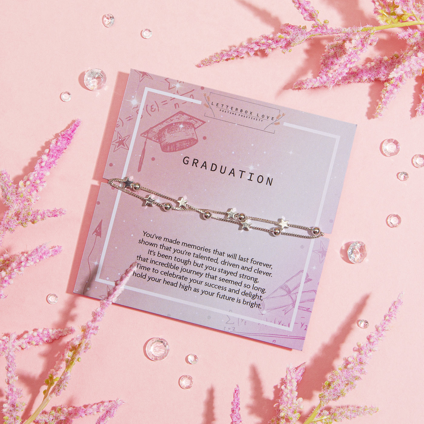 GRADUATION' bracelet with star charms and a solitary pearl, displayed on a soft pink card adorned with illustrations of a graduation cap, arrows, and math symbols. The card carries an uplifting message celebrating a graduate's achievements and bright future.