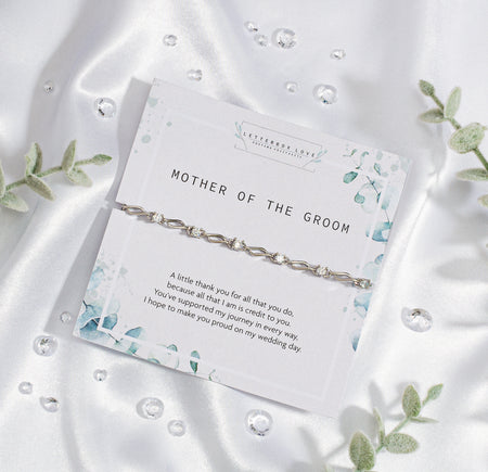 Elegant 'Mother of the Groom' card set against a silky white fabric backdrop. The card, adorned with soft blue floral designs, expresses gratitude and appreciation with a heartfelt message. Draped over the card is a sophisticated silver bracelet with intricate detailing. 