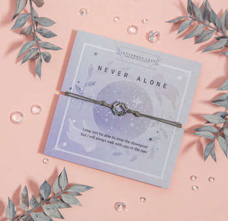 NEVER ALONE' bracelet with a square-shaped gemstone charm, presented on a pale blue card adorned with celestial designs and a reassuring message about companionship during tough times. 