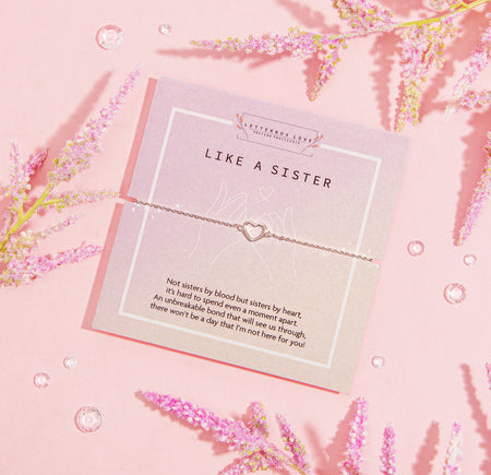 LIKE A SISTER' bracelet featuring a heart charm, presented on a muted pink card with a heartfelt message about the deep bond of friendship that's akin to sisterhood. The card is adorned with delicate pink blossoms and clear crystal gems, set against a soft pink backdrop