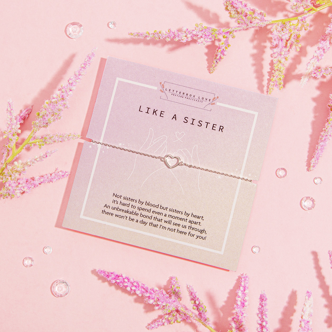 LIKE A SISTER' bracelet featuring a heart charm, presented on a muted pink card with a heartfelt message about the deep bond of friendship that's akin to sisterhood. The card is adorned with delicate pink blossoms and clear crystal gems, set against a soft pink backdrop