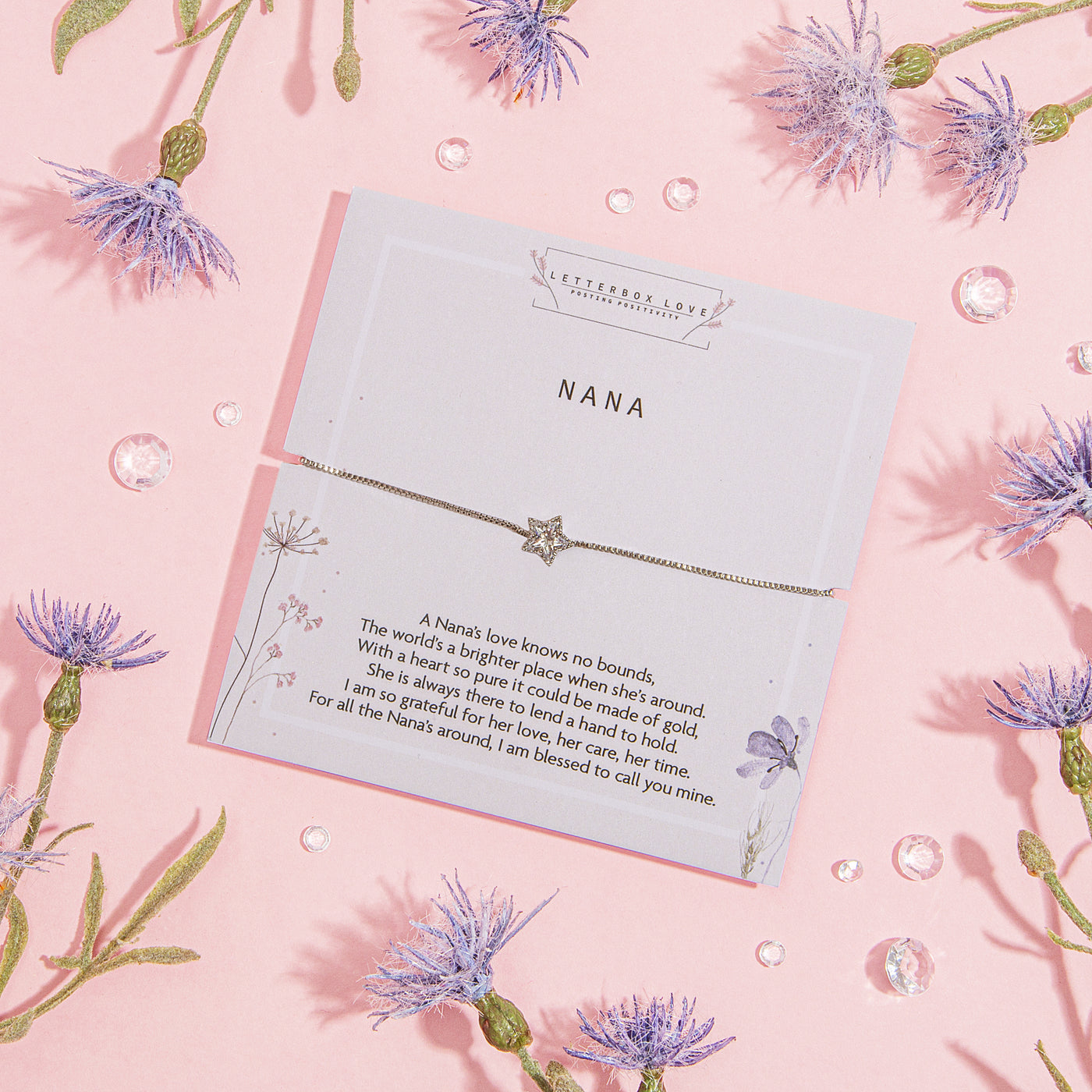 Special card titled 'NANA' with a heartfelt message celebrating a Nana's love, complemented by an elegant bracelet featuring a star charm. The card is presented on a soft pink background, decorated with sparkling droplets and vivid blue thistle-like blossoms.