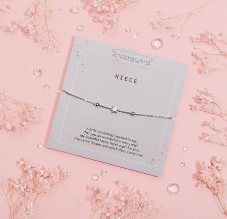Pastel pink greeting card titled 'NIECE' with heartfelt message, adorned with silver star motifs, accompanied by a dainty silver bracelet featuring a star charm, surrounded by delicate white baby's breath flowers and clear gemstones.