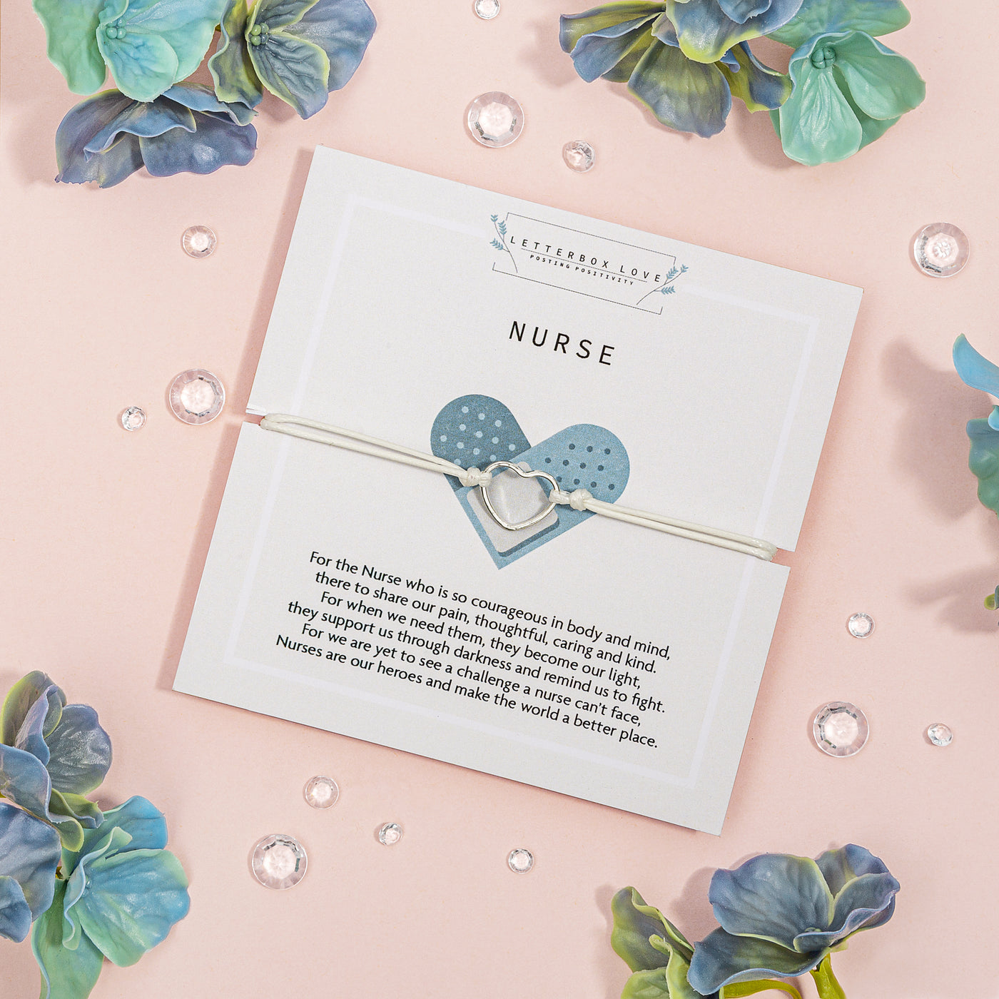 White bracelet with a silver heart-shaped charm showcased on a white 'NURSE' appreciation card with a heartfelt message about their courage and care, surrounded by blue-green flowers and crystal accents on a soft pink background