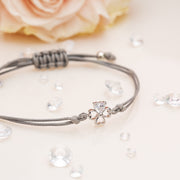 The image shows a delicate bracelet placed on a soft, light-colored surface with a blurred pink rose in the background. The bracelet features a silver four-leaf clover charm with a sparkling heart-shaped gem at its center, attached to a grey cord. The cord is tied in an adjustable knot design for easy resizing. Scattered around the bracelet are small, clear decorative gems, enhancing the elegant and charming aesthetic of the piece.